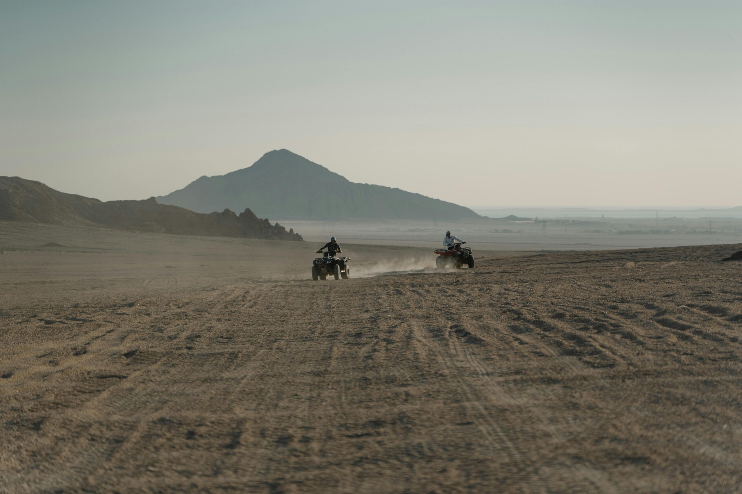 Men riding ATVs across a dusty desert path with mountainous backdrop, creating a thrilling adventure scene.